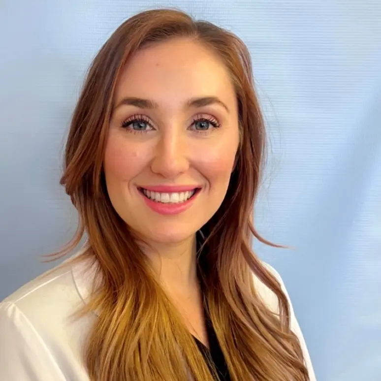 A woman with long light brown hair and a white top smiles at the camera against a plain background.