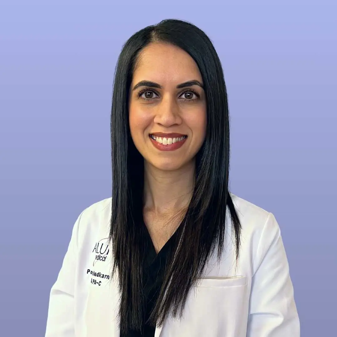 A person with long black hair wearing a white lab coat and black shirt smiles against a light purple background.