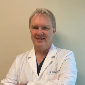 A man in a white lab coat stands against a beige wall with his arms crossed. His name is embroidered on his coat.